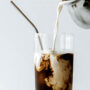 tall glass of iced CBD infused coffee with a metal straw in the glass; milk is being poured into the iced coffee glass