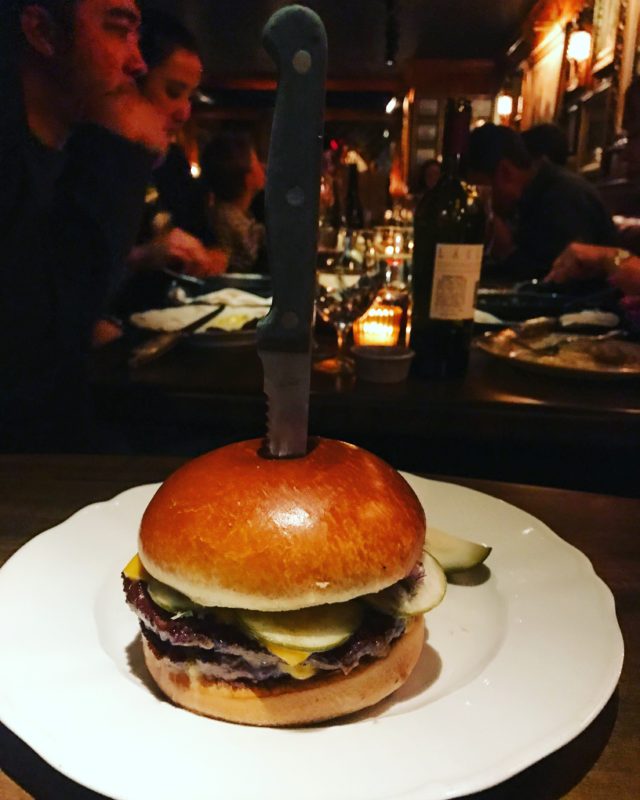 round bun; double patty 4 Charles Prime Rib cheeseburger with steak knife right in the middle; sliced pickle behind burger