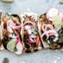 artisan photo of five chicken tacos in charred white soft taco bread; garnished with radish;red onion; and feta cheese with parsley and a sliced lime