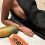 tanned woman in black jumpsuit showing side boob while eating fruit on beach