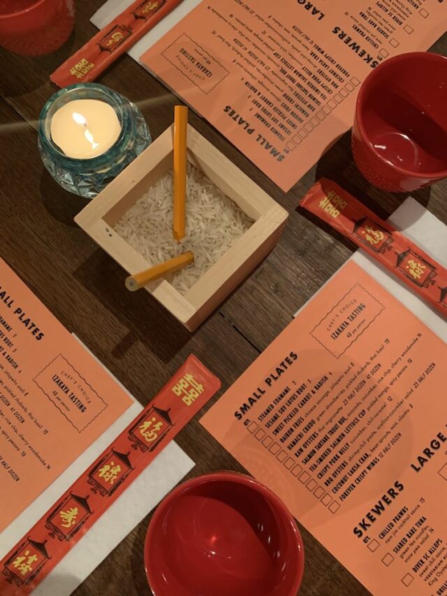 The Poni Room’s orange menu on a brown wooden table; An Izakaya Bar table with a rice box and two pencils in the center 