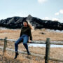 Monica Dimperio sitting on a wooden fence with a fun smile; she has long blonde hair tied back by a black and white head tie; her hair is flowing in the wind; she is wearing an olive patagonia fleece with blue jeans and black chelsea boots; the background shows the mountains of Boulder; the mountains are gray colored with green pine trees and little bit of snow